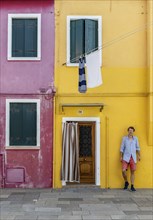 Young man in front of a house wall, red and yellow house facade with entrance door and windows,