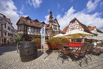 Market fountain with geraniums, tables, chairs and parasols of a restaurant and half-timbered