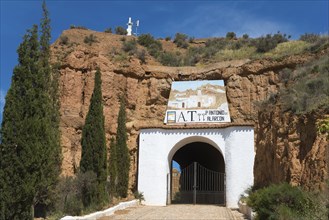 Entrance gate to a white building with a rock face and trees, surrounded by rustic charm on a sunny