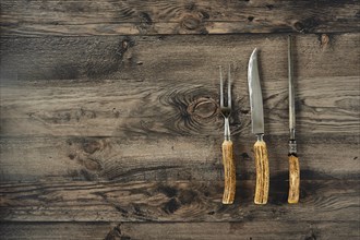 Three essential barbecue tools on a rustic kitchen table made of dark wood, knife, fork and