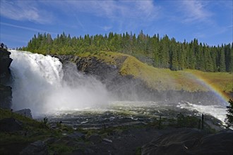 A large waterfall cascades thunderously into a river, a rainbow is visible, surrounded by dense