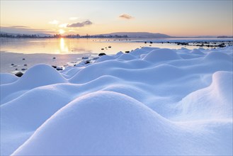 Gentle hills of snow stretch towards the lake, where the sun sets over the mountains, Seegarten,