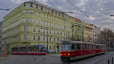 Several generations of trams run on cobblestone streets with old buildings in Prague, overcast sky,