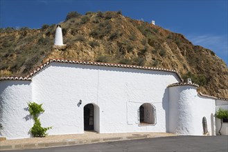 White-painted building with chimney and windows in front of a rock face, under a sunny blue sky,