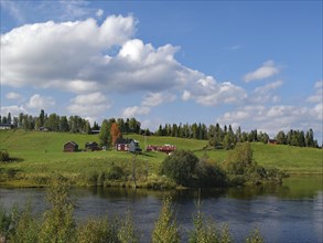 A river flows along a green meadow with farmhouses, the sky is blue, some clouds, autumn, Störlien,