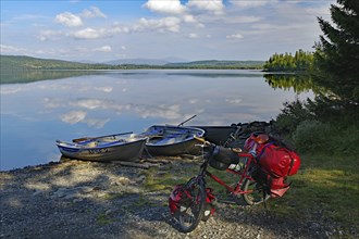 A lake with docked boats, a bicycle standing on the shore, surrounded by trees and under a cloudy