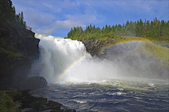 A large waterfall flows over rocks while a rainbow can be seen at the water's edge and forest