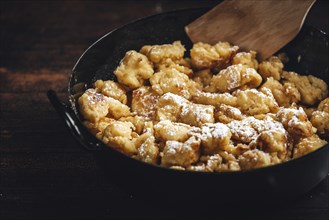 Kaiserschmarrn in a pan dusted with sugar and wooden spatula, close-up, dark background