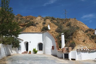 Small white building with chimney, tree and rock face in the background under a sunny blue sky,