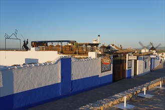 Rustic building with white and blue walls, tapas signs and windmills in the background under a