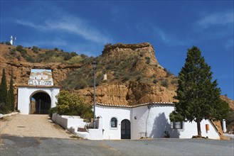 White building with entrance gate and trees in front of a rock face, under a sunny, cloudy sky,