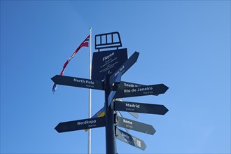 Black signpost in front of a blue sky and a Norwegian flag, with directions to various cities and