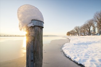 A snow-covered wooden post at the edge of a frozen body of water, trees in the background,