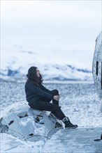 A woman sits on the engine of a crashed plane in the frozen Solheimasandur square in winter in