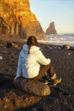 Female hiker sitting on the black sand beach of Reynisfjara looking at the sea. Iceland