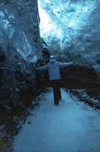 A man is walking through a cave with a blue sky above him. He is wearing a blue jacket and black