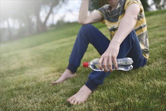 Young caucasian man runner relaxing holding drinking water bottle and sitting on grass in the park