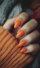 A close-up of a hand with vibrant orange nail polish and wearing a warm orange sweater creating a