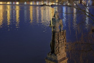 Statue of a knight with a sword overlooking a river with reflections of lights on the water at