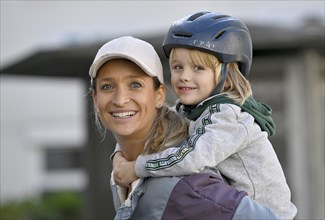 Mother, woman, blond, baseball cap, smiling, wearing boy with helmet, 5 years, blond, on shoulder,