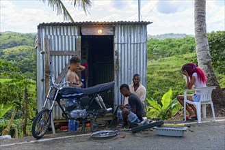 A group of people repairing a motorbike in front of a metal hut surrounded by green landscape,