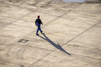 Man, labourer, with work suit, hard hat, walking across a depot, shadow