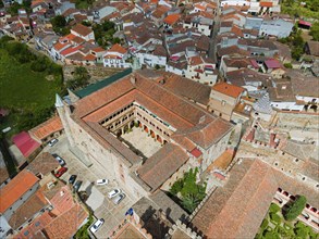 Aerial view of a monastery with a central courtyard and red roofs, surrounded by a village.