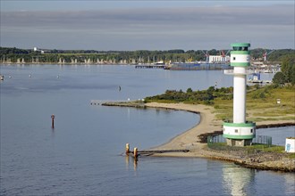 White-green lighthouse on a coast with calm water, in the background boats and a harbour,