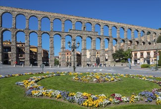 A historic stone aqueduct with arches spans a city under a bright blue sky next to a colourful