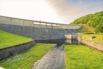 A mighty concrete dam with a river in the foreground, surrounded by green land and trees,