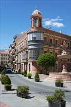 Historic building with bay window on an urban street with fountain and trees under a blue sky, Casa