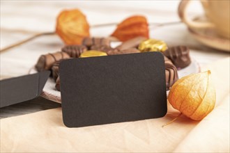 Black business card with chocolate candies and physalis flowers on a white wooden background and