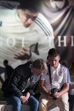 Two visitors sitting in front of a banner looking at a mobile phone at the Adidas fan zone at the