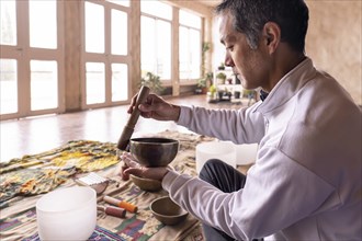 A middle-aged Asian man sits in profile, concentrating on gently vibrating a brass bowl in a softly