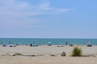 Beach with people and colourful umbrellas, stones and grass, in front of a wide, blue Mediterranean