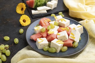 Vegetarian salad with watermelon, feta cheese, and grapes on blue ceramic plate on black concrete