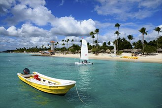 A sunny beach with clear blue water and a boat in the foreground, lighthouse, bar of the Hotel