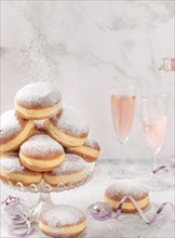 Doughnuts on a glass stand, rosé sparkling wine is poured in the background, falling icing sugar