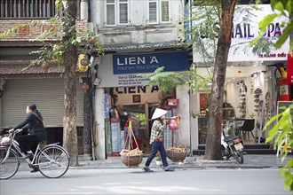 Vietnamese saleswoman in a conical hat shoulders cassava on a carrying pole through the Old Quarter