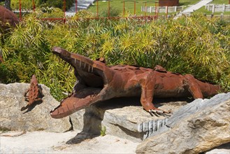 Metal sculpture of a crocodile with open mouth on a rock foundation surrounded by plants in the