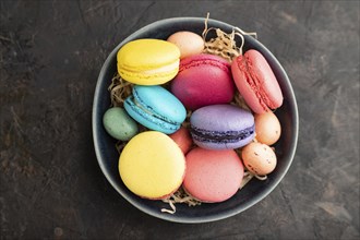 Multicolored macaroons and chocolate eggs in ceramic bowl on black concrete background. top view,