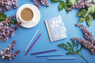 Purple lilac flowers and a cup of coffee with notebook and colored pencils on pastel blue