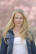 Woman with blonde hair wearing a denim jacket and white top smiles with a soft pink background in