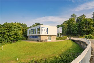 Modern, white building on a green meadow under a blue sky next to a bridge, Rappbodetalsperre, Harz