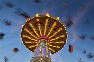 An illuminated carousel at dusk, spinning fast, in front of a blue sky with clouds and a Germany