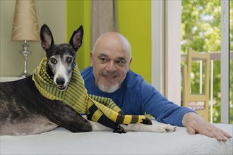 Portrait of a man posing and looking at his rescued greyhound dog with a green scarf lying on the