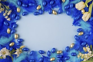 Frame of blue and turquoise feathers with blue and gold chocolate eggs, spring flowers, on a light