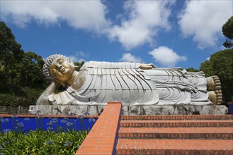 Reclining Buddha statue above an area decorated with flowers and steps in a sunny park, Bacalhôa,