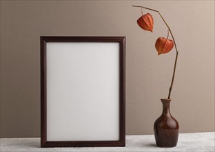 Brown wooden frame mockup with physalis in ceramic vase on beige paper background. Blank, vertical