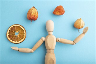 Wooden mannequin juggling dried citrus and physalis on blue pastel background. close up, isolated